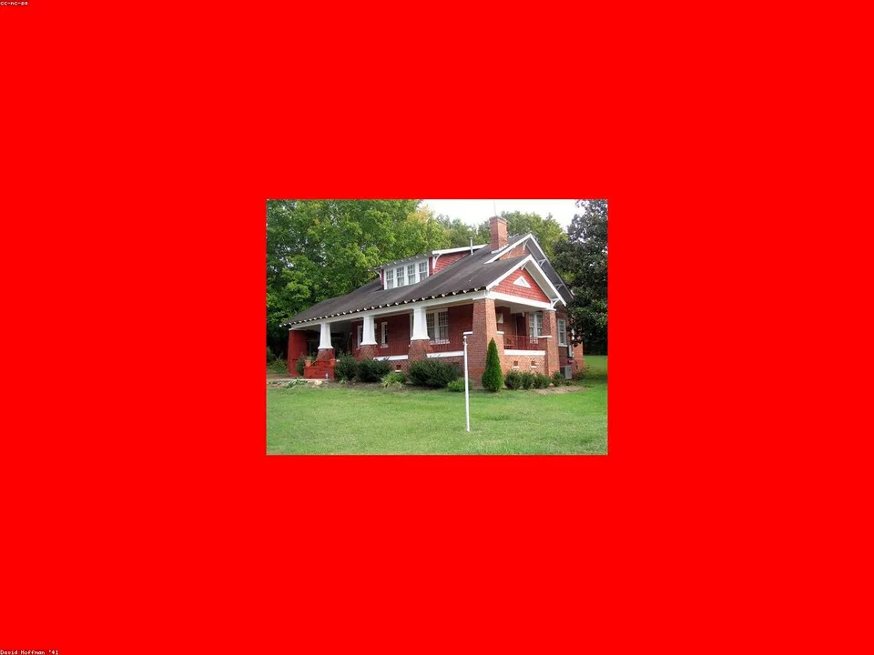 Red brick single-story house with a pitched roof and front porch, surrounded by green lawn and trees.