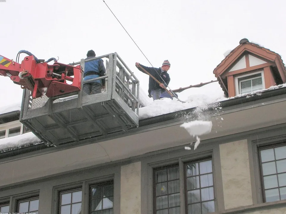 Two workers on a lift-elevated platform clearing heavy snow from a building's roof edge, with a red brick chimney visible on the right side.
